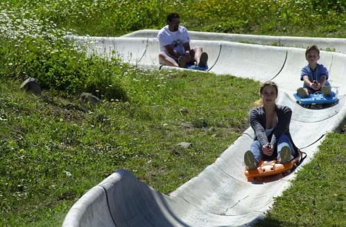 A family enjoys a summer day sliding down a grassy hill on sleds, surrounded by flowers and greenery.