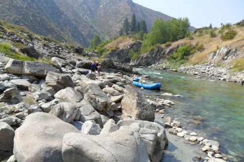 A rocky riverbank with a blue raft on the water, surrounded by mountains and greenery under a clear sky.
