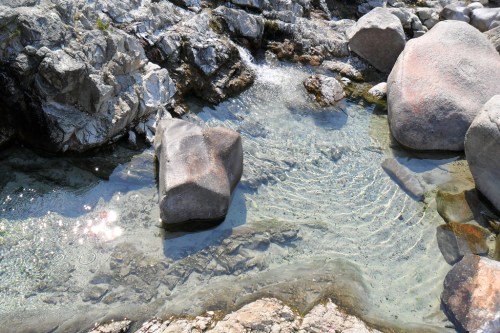 Clear water flows over smooth rocks in a serene natural setting, with sunlight reflecting on the surface.