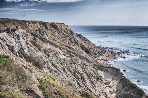 A rugged coastline with steep cliffs overlooking a calm ocean under a cloudy sky.