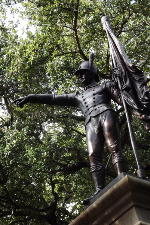 A bronze statue of a soldier holding a flag, pointing forward, surrounded by lush green trees.