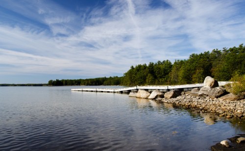 A serene lake scene with a wooden dock, surrounded by trees and rocky shoreline under a blue sky with wispy clouds.