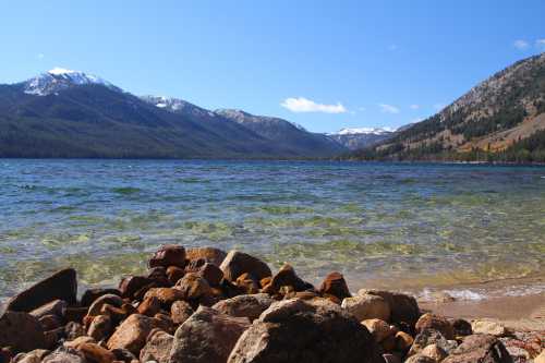 A serene lake surrounded by mountains, with clear water and rocky shore under a blue sky.