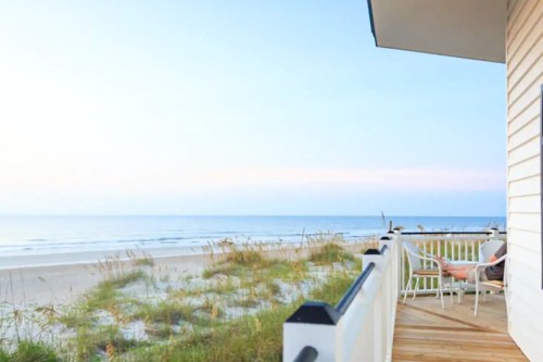 A serene beach view from a balcony, with gentle waves and grass in the foreground under a clear sky.