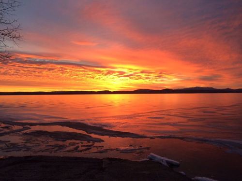 A vibrant sunset over a calm lake, with colorful clouds reflecting on the water and ice along the shore.
