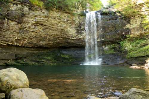 A serene waterfall cascades into a clear pool, surrounded by lush greenery and rocky cliffs.