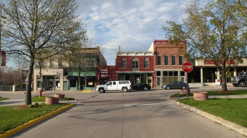 A street view of historic buildings with trees, a stop sign, and a white truck parked in the foreground.