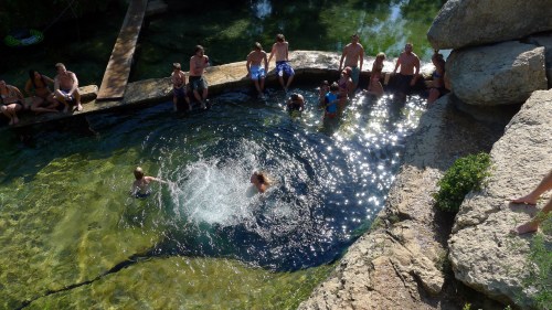 A sunny outdoor scene with people swimming and relaxing by a clear water pool surrounded by rocks.