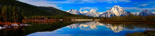 Scenic mountain landscape with snow-capped peaks reflecting in a calm lake, surrounded by lush forests and blue sky.
