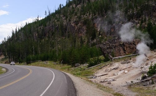 A winding road alongside a forested hillside with steam rising from the ground, indicating geothermal activity.
