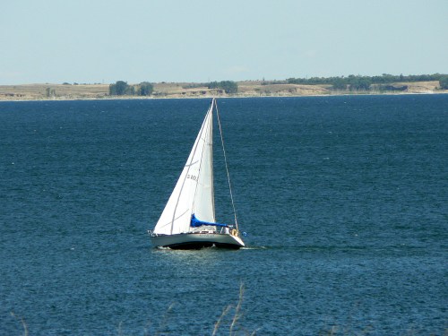 A sailboat with a white sail glides across a calm blue lake under a clear sky.