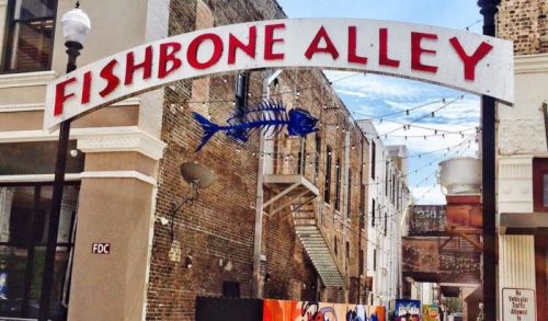 Sign for Fishbone Alley with a fish skeleton graphic, surrounded by brick buildings and string lights.