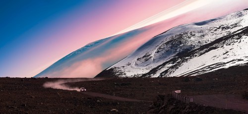 A car drives on a winding road through a rocky landscape, with snow-capped mountains and a colorful sky at dusk.