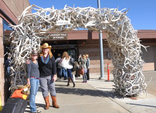 Two women pose under a large antler archway at Jackson Hole, Wyoming, with people walking in the background.