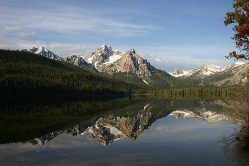 A serene lake reflects snow-capped mountains and lush green forests under a clear blue sky.