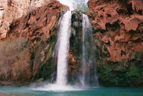 A stunning waterfall cascades over red rock cliffs into a turquoise pool, surrounded by lush greenery.