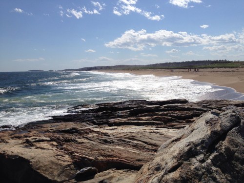 A rocky shoreline with waves crashing, leading to a sandy beach under a blue sky with scattered clouds.
