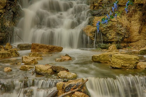 A serene waterfall cascades over rocks, surrounded by lush greenery and blue flowers.