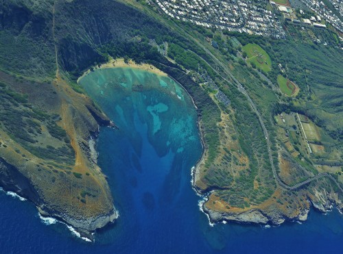 Aerial view of a coastal bay with clear blue water, surrounded by green hills and a nearby residential area.