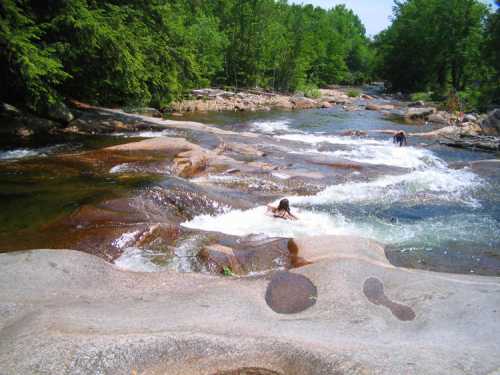 A serene river flows over smooth rocks, with lush green trees lining the banks and two people enjoying the water.