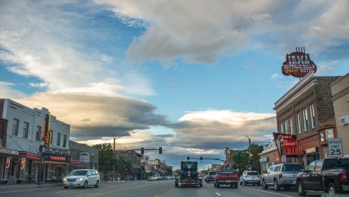 A street view of a small town with shops, a theater, and a cloudy sky at sunset. Cars are driving along the road.