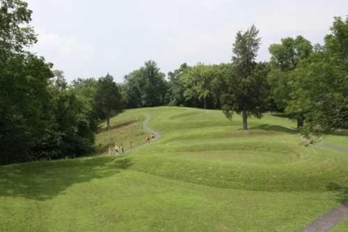 A winding path through grassy hills, with trees and a few people walking in the distance under a cloudy sky.