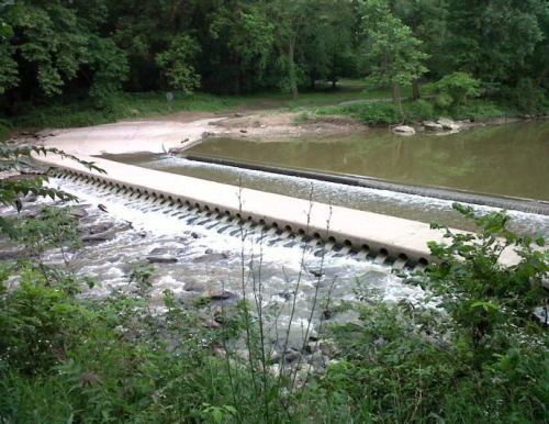 A concrete dam with water flowing over it, surrounded by lush greenery and a calm river.