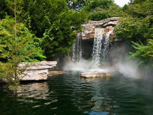A serene waterfall cascades into a misty pond, surrounded by lush greenery and rocky formations.