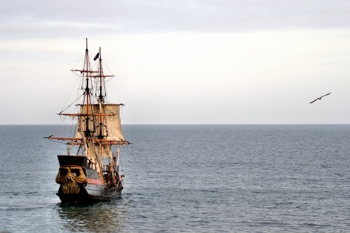 A pirate ship with a skull figurehead sails on calm waters under a cloudy sky, with a bird flying nearby.