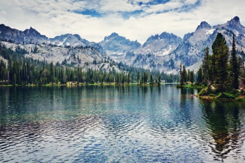 A serene lake surrounded by mountains and lush trees under a cloudy sky. Reflections shimmer on the water's surface.