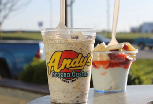 A cup of frozen custard from Andy's, with a spoon, next to a parfait of fruit and yogurt on a table.