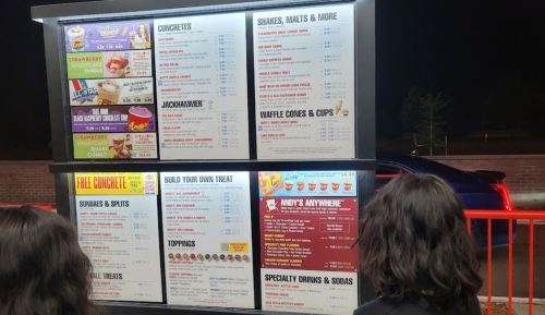 Two people stand in front of a brightly lit menu board at a dessert shop, reviewing various treats and options.
