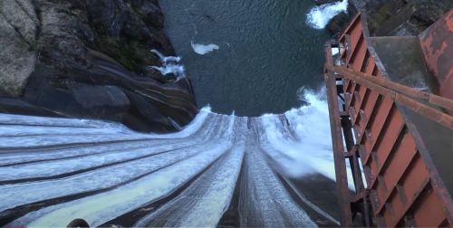 Aerial view of a large dam with water cascading down into a river below, surrounded by rocky terrain.