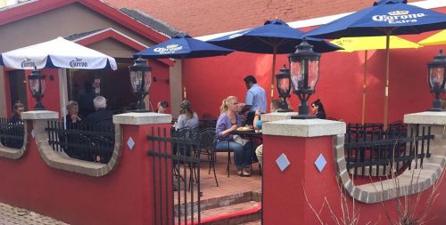 Outdoor dining area with umbrellas, people seated at tables, and a vibrant red wall in the background.