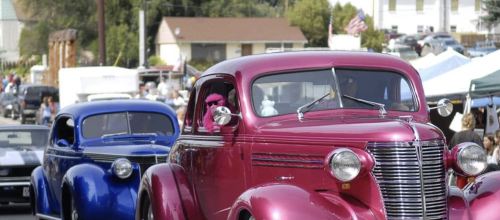 A parade featuring classic cars, including a pink vintage car and a blue one, with people and tents in the background.
