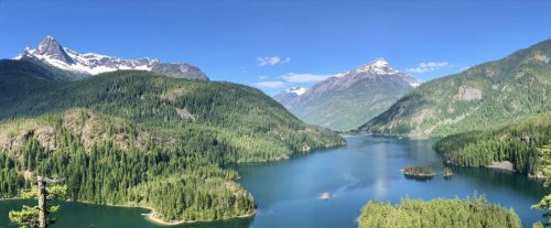 A panoramic view of a serene lake surrounded by lush green forests and majestic mountains under a clear blue sky.