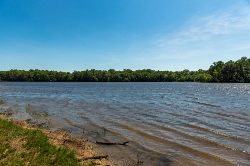 A calm river under a clear blue sky, bordered by lush green trees along the shoreline.