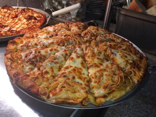 A large pizza topped with melted cheese and spaghetti, sliced into triangular pieces on a metal tray.