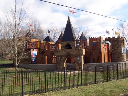 A wooden castle play structure with turrets and flags, surrounded by grass and a black fence.