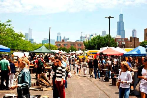 A bustling outdoor market with colorful tents, shoppers browsing, and a city skyline in the background.