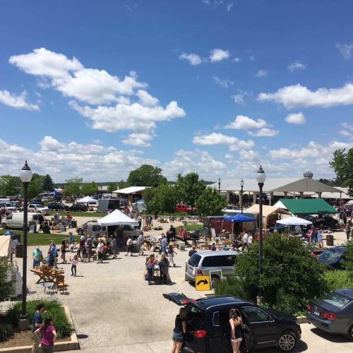 A bustling outdoor market scene with tents, vendors, and crowds under a blue sky with fluffy clouds.