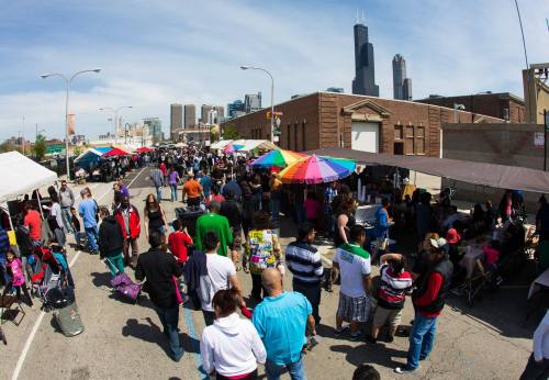 A bustling street market with colorful tents, crowds of people, and city skyscrapers in the background on a sunny day.