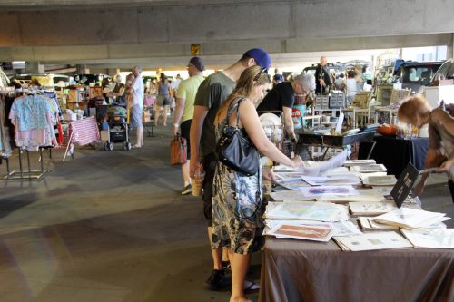A bustling market scene with people browsing tables of art and crafts under a parking structure.