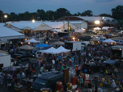 A bustling outdoor market at dusk, filled with people, tents, and various vendors under bright lights.