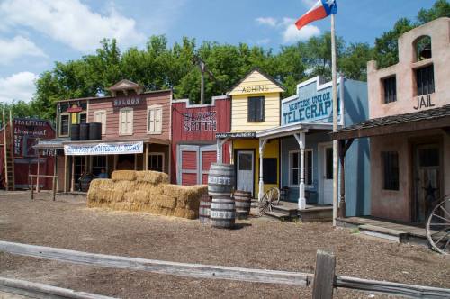 A colorful old western town with wooden buildings, hay bales, and a Texas flag in the background.