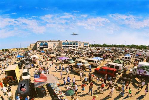 A bustling outdoor market with numerous stalls, vehicles, and people, set against a clear blue sky and an airplane overhead.