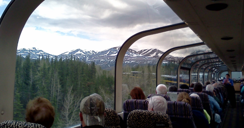 Passengers sit inside a train, gazing at snow-capped mountains and lush forests through large panoramic windows.