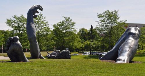 Large bronze sculptures of a hand and foot emerging from the ground, surrounded by green grass and trees.