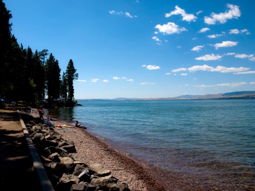 A serene lakeside scene with clear water, rocky shore, and trees under a bright blue sky with scattered clouds.