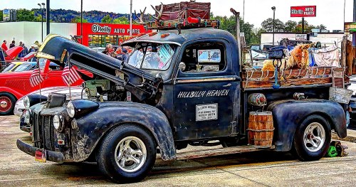 A vintage truck with an open hood, decorated with flags and a dog, parked at a car show. "Hillbilly Heaven" is painted on the side.